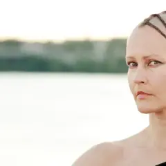 Virpi Pähkinen med flätor på rakad skalle, blickar mot betraktaren vid en sjö.