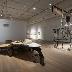 Konstinstallation med en stor träkonstruktion och en skulptur av metall och ben. En accelerator su-liknande enhet hänger från taket.