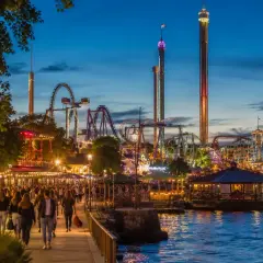 Kväll vid Gröna Lund med folk som promenerar längs vattnet, berg-och-dalbanor och höga torn som lyser upp himlen.