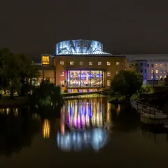 Nattbild över De Geerhallen där ett evenemang pågår, med färgade ljus som reflekteras i vattnet.