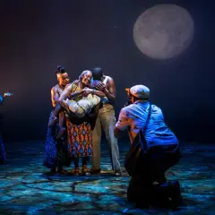 Scen från en teaterföreställning med en grupp skådespelare i färggranna kläder, en stor måne lyser upp scenen. En fotograf fångar ögonblicket. En påminnelse om alla fantastiska teatrar i Stockholm.