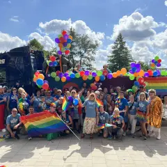 Stor grupp människor firar med regnbågsflaggor och ballonger vid en pride park.