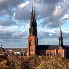 Uppsala domkyrka med sina höga torn reser sig mot en dramatisk molnhimmel. Höstlöven lyser i gult och orange.