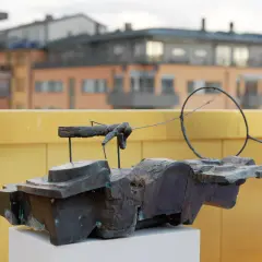En abstrakt skulptur i mörk metall, placerad på ett vitt podium. I bakgrunden syns byggnader och en gul vägg, kanske vid museum Vasaparken.