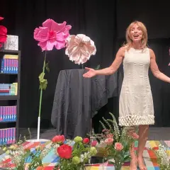 En kvinna talar på en scen med stora pappersblommor och bokhyllor, en del av Rinkeby bokfestival.