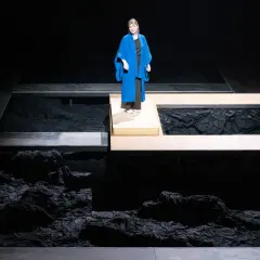 En kvinna i blå kappa står på en upphöjd plattform under en föreställning av Hamlet i Stockholm.