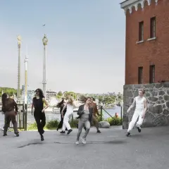 Dansare utför en koreografi utomhus, med en tegelbyggnad och nöjespark i bakgrunden. En scen som påminner om moderna dansteatern.