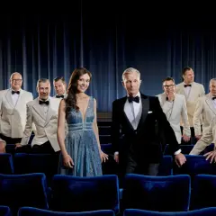 Max Raabe och hans Palast Orchester poserar i vita kostymer och en glittrig klänning framför en mörkblå ridå, redo för en föreställning i Stockholm.