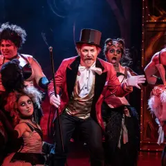 Scenen från Moulin Rouge! visar en cirkusdirektör med en cylinderhatt och en gyllene väst, omgiven av artister i glittrande kostymer. Handlar om kärlek och frihet i Paris.