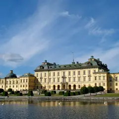Drottningholm slott, ett gult barockpalats med gröna tak, speglar sig i vattnet under en blå himmel med tunna moln.