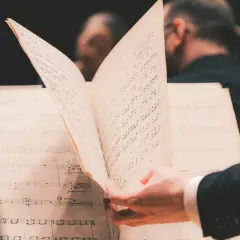 En hand bläddrar i noter under en lunchkonsert på operan. Musiker i bakgrunden förbereder sig.