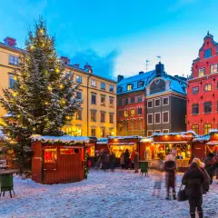 En snötäckt vinterviken julmarknad med ett stort, upplyst träd och färgglada hus. Folk strosar bland bodarna.