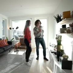 Två personer diskuterar olika inredningsstilar i ett ljust vardagsrum med modern möblering, en mörk soffa och hyllor med växter.