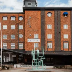 Magasin III, en byggnad i tegel med många fönster, har en flagga med texten "Magasin III". Framför finns en grön skulptur.