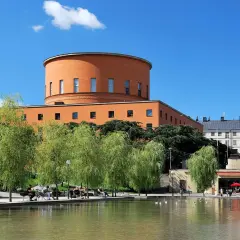 En vacker stadsvandring i Stockholm på egen hand. En orange, rund byggnad reser sig bakom grönskande träd vid en damm.