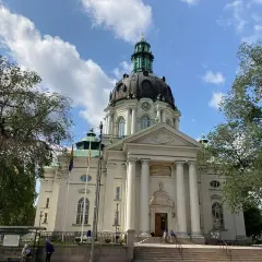 Imponerande Gustav Vasa kyrka med sin kupol och pelare, omgiven av träd och en blå himmel med moln.