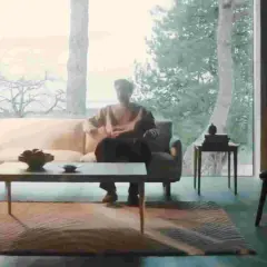 En man sitter i en soffa i ett ljust rum med stora fönster. En dansk designer kan ha skapat den moderna inredningen.