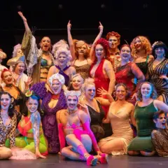 En stor grupp artister i glittrande kostymer poserar glatt på scenen under Stockholm Burlesque Festival.