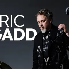 Eric Gadd, med sitt karaktäristiska lockiga hår och skägg, står redo att uppträda. En gitarr och mikrofon antyder en musikalisk kväll på Mosebacke.