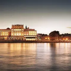 Kungliga operan i Stockholm lyser upp vid vattnet i skymningen, dess fasad speglas i det lugna vattnet.
