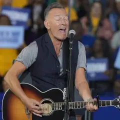 Bruce Springsteen sjunger och spelar gitarr på scenen. Publiken håller upp skyltar, vilket antyder ett kommande evenemang med Bruce Springsteen.