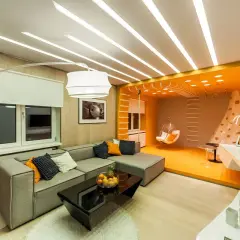 Stort vardagsrum med en grå soffa, svart soffbord och en orange sittgunga. Taket har en unik design med många ljuslister.