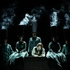 Scen från Sweeney Todd operan. Människor med höga hattar och rök som stiger ur dem. En man skriver vid ett bord.
