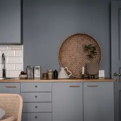 Ett kök med gråa skåp och en träbänk. En rund flätad bricka med en växt, en vit kanna och två metallburkar skapar ett vackert stilleben inredning.