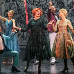 Scen från "Tootsie" på Oscarsteatern. En man i ljusblå kostym knäböjer medan en kvinna i glittrig grön klänning och andra i färgglada klänningar dansar.