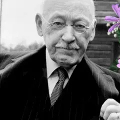 Äldre man med glasögon och mustasch, klädd i kostym. Bredvid honom en bukett med lila och gula blommor. Han är alfven som skrev.