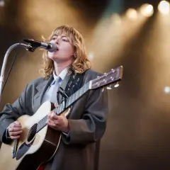 Ellen Krauss konsert. En artist med blont hår, klädd i kavaj och slips, spelar gitarr och sjunger i mikrofonen på en scen med strålkastare.
