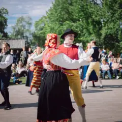 Dansande par i folkdräkter på en utescen, publiken tittar på. En härlig konsert på Skansen.