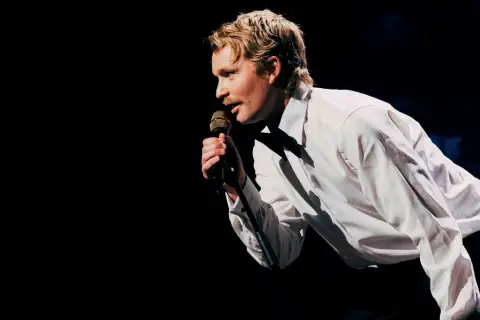 En man med mustasch, klädd i vit skjorta, sjunger i mikrofonen under nalles show.