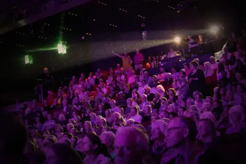 Publik njuter av en konsert i Uppsala. Scenen badar i lila ljus, och en artist gestikulerar mot publiken.