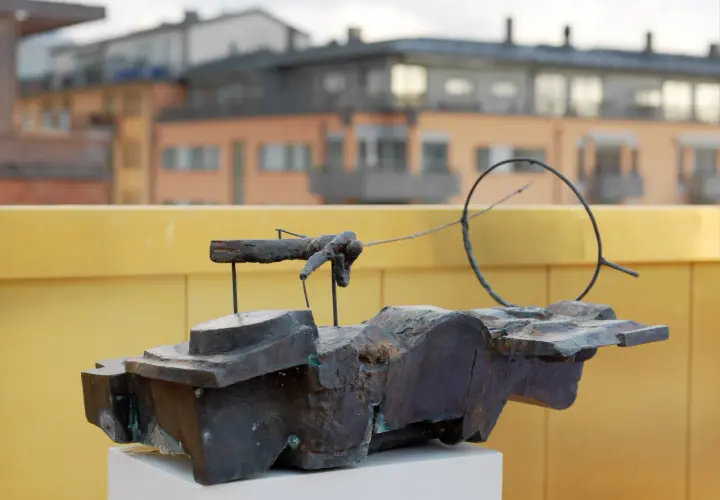 En abstrakt skulptur i mörk metall, placerad på ett vitt podium. I bakgrunden syns byggnader och en gul vägg, kanske vid museum Vasaparken.
