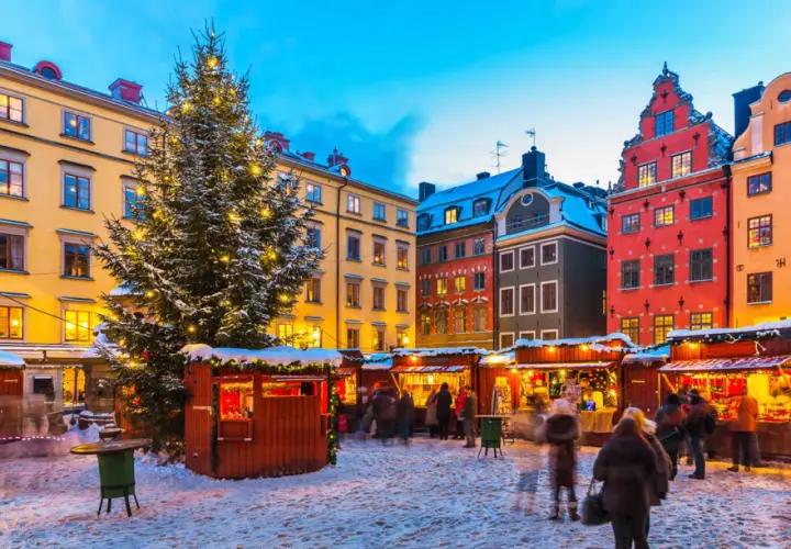 En snötäckt vinterviken julmarknad med ett stort, upplyst träd och färgglada hus. Folk strosar bland bodarna.