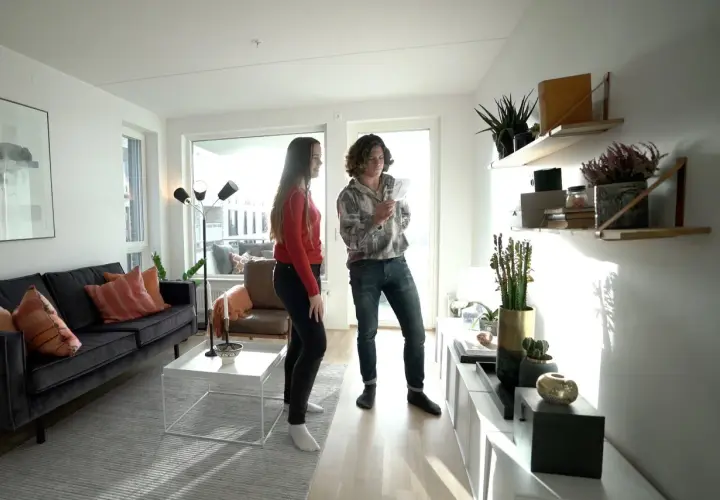 Två personer diskuterar olika inredningsstilar i ett ljust vardagsrum med modern möblering, en mörk soffa och hyllor med växter.