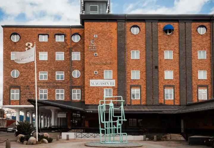 Magasin III, en byggnad i tegel med många fönster, har en flagga med texten "Magasin III". Framför finns en grön skulptur.
