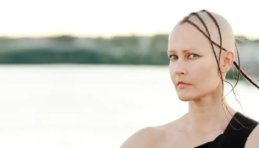 Virpi Pähkinen med flätor på rakad skalle, blickar mot betraktaren vid en sjö.