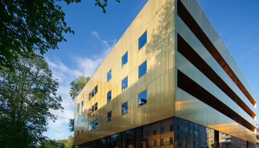 Guldhuset Vasaparken glänser i solen med sin gyllene fasad och stora fönster. Trädens grönska ramar in den moderna byggnaden.