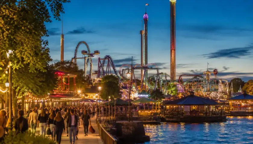 Kväll vid Gröna Lund med folk som promenerar längs vattnet, berg-och-dalbanor och höga torn som lyser upp himlen.