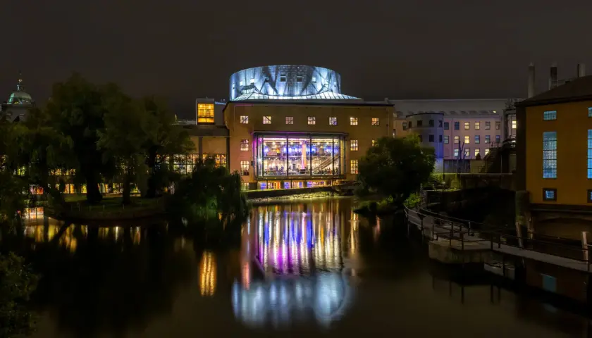Nattbild över De Geerhallen där ett evenemang pågår, med färgade ljus som reflekteras i vattnet.