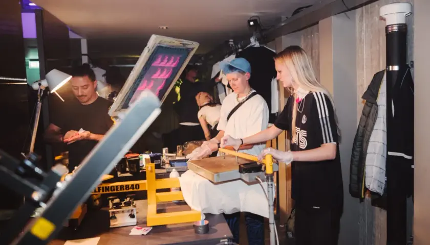 Människor arbetar med screentryck i ett kreativt hus. En man förbereder sig, medan två kvinnor hanterar tryckpressen.
