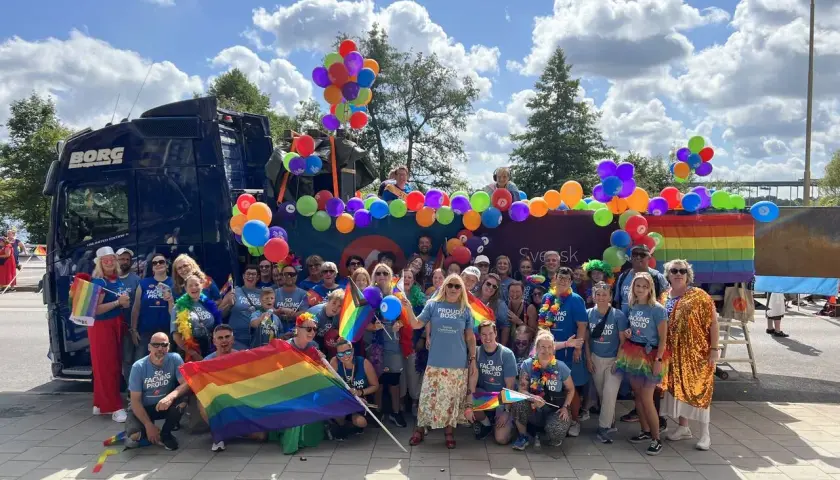 Stor grupp människor firar med regnbågsflaggor och ballonger vid en pride park.
