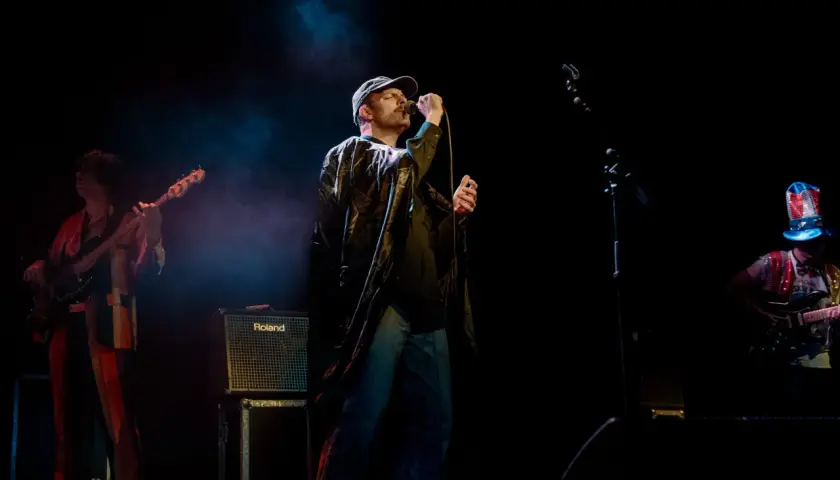 Mac DeMarco sjunger på scen i Stockholm, iklädd keps och en glansig jacka. En basist och en gitarrist syns också.