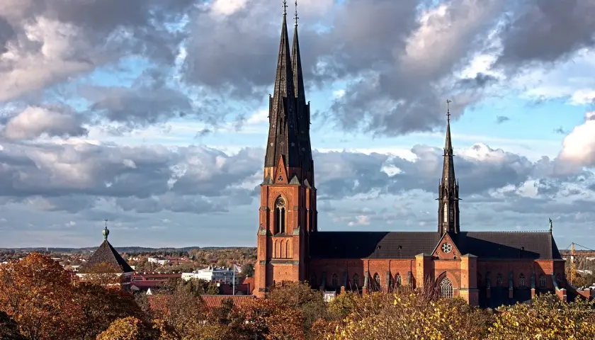 Uppsala domkyrka med sina höga torn reser sig mot en dramatisk molnhimmel. Höstlöven lyser i gult och orange.