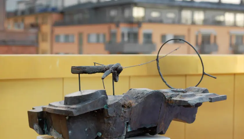 En abstrakt skulptur i mörk metall, placerad på ett vitt podium. I bakgrunden syns byggnader och en gul vägg, kanske vid museum Vasaparken.