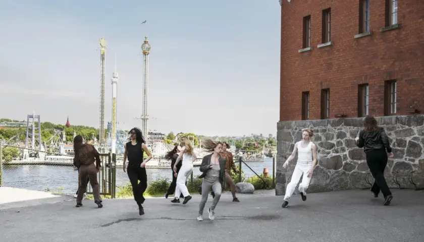 Dansare utför en koreografi utomhus, med en tegelbyggnad och nöjespark i bakgrunden. En scen som påminner om moderna dansteatern.