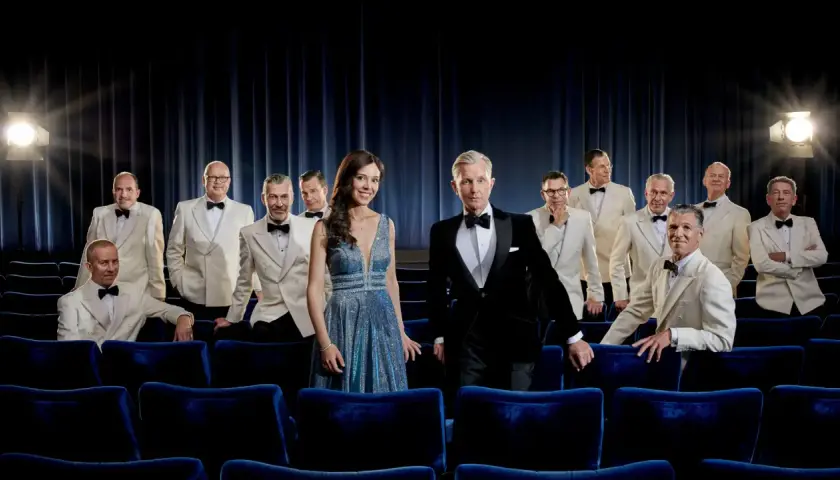 Max Raabe och hans Palast Orchester poserar i vita kostymer och en glittrig klänning framför en mörkblå ridå, redo för en föreställning i Stockholm.