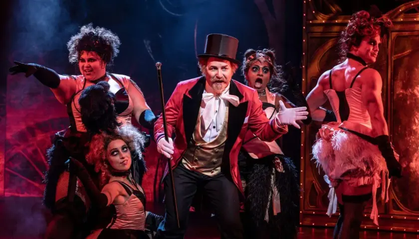 Scenen från Moulin Rouge! visar en cirkusdirektör med en cylinderhatt och en gyllene väst, omgiven av artister i glittrande kostymer. Handlar om kärlek och frihet i Paris.